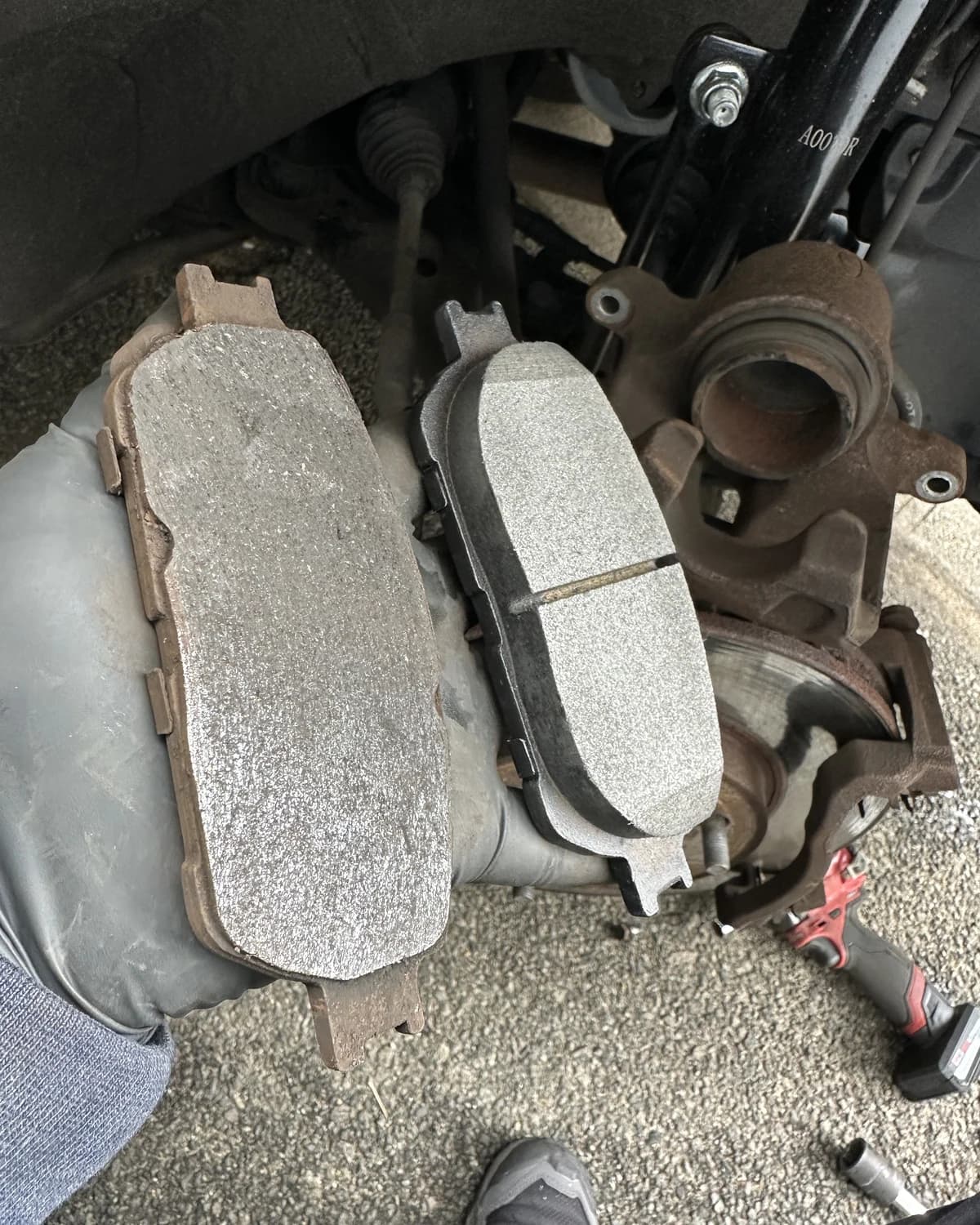 A gloved hand comparing a used brake pad with a new brake pad beside an exposed brake assembly.