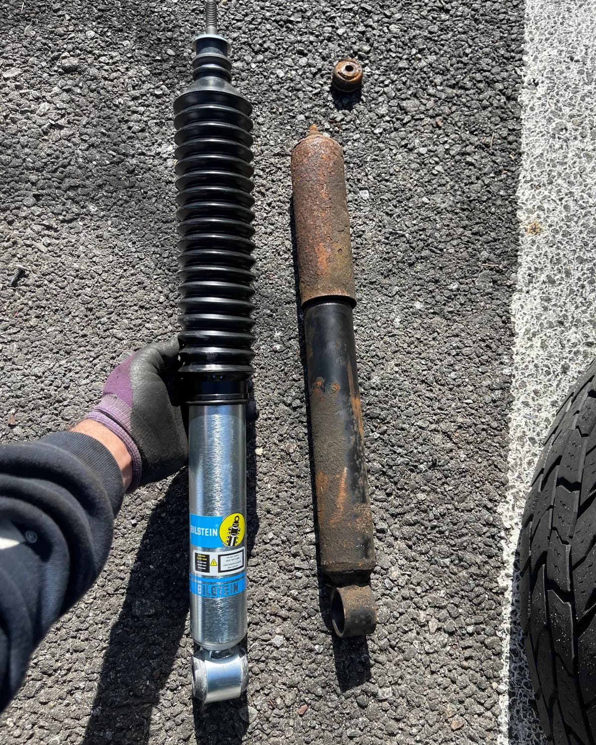 A gloved hand comparing a new shock absorber with a worn, rusted shock absorber.
